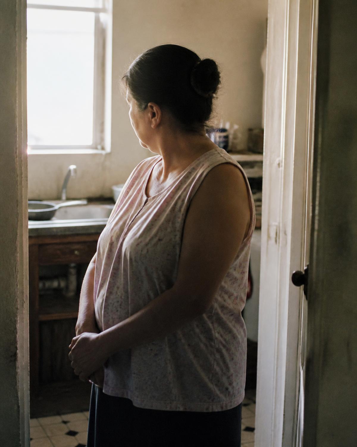 A woman pausing in a modest kitchen doorway, looking away from the camera