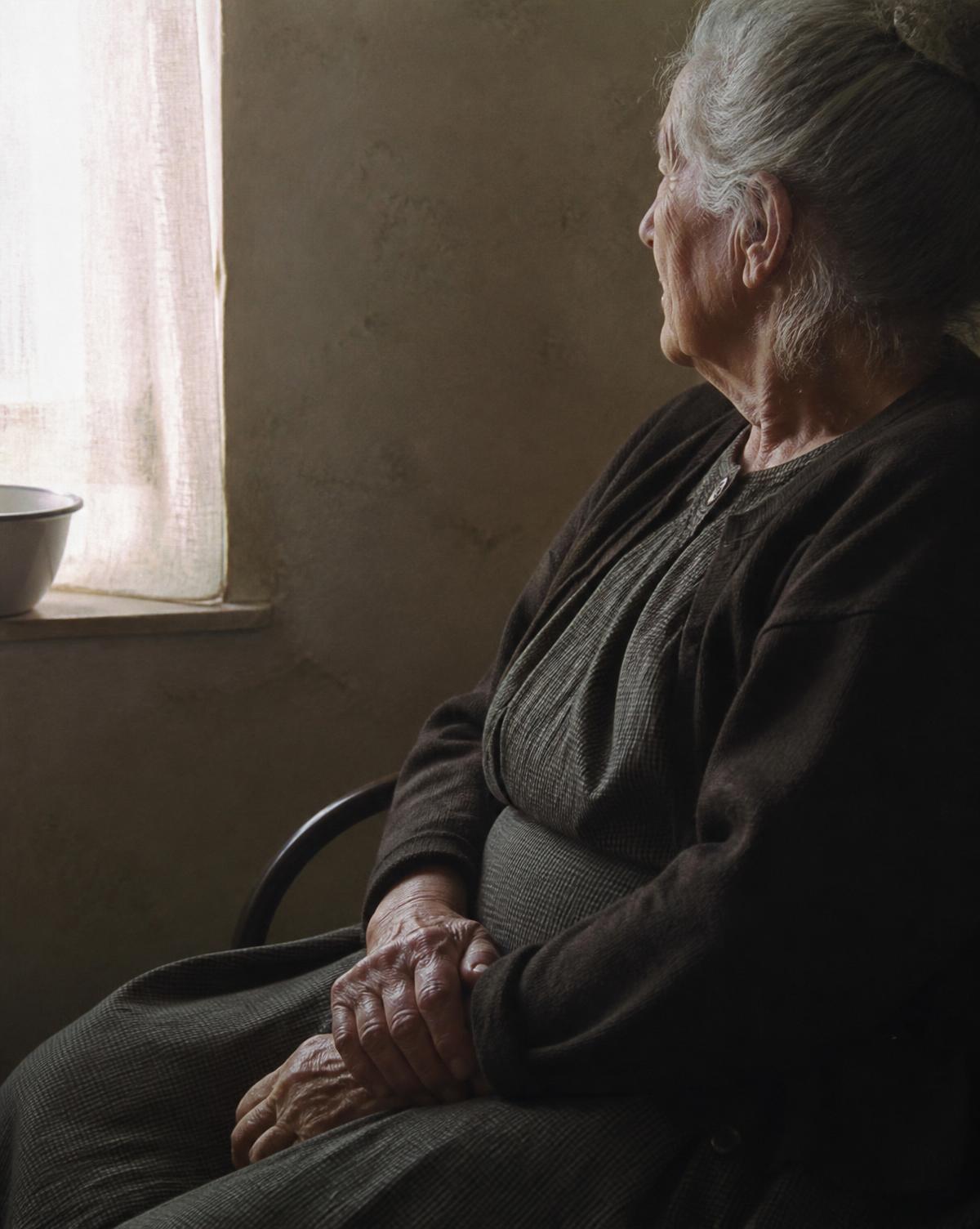An elderly woman seated by a window, hands folded, turned toward the light