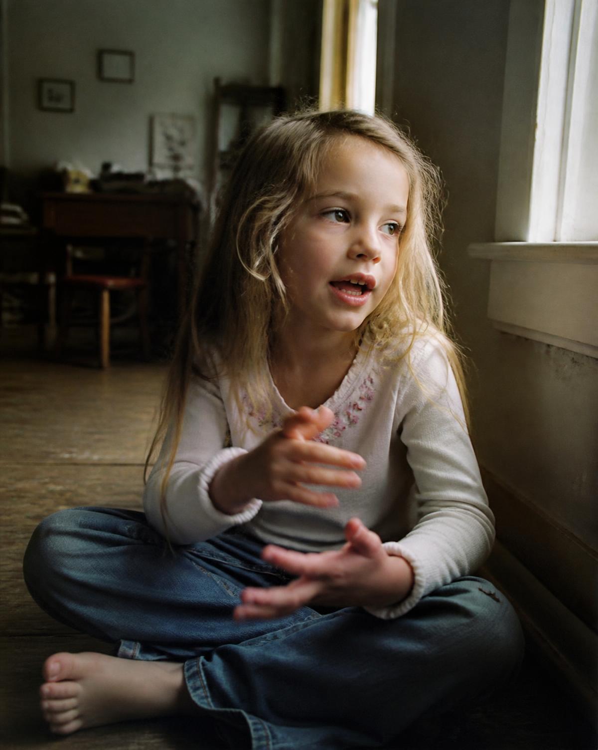 A young child sitting on a wooden floor near a window, mid-sentence, hands moving